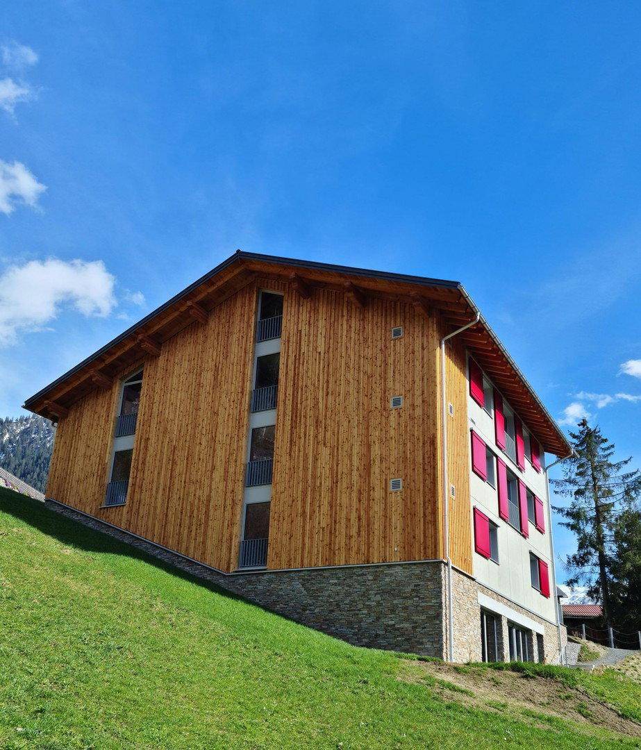 Ansicht des modernen Gebäudes mit Holzfassade und roten Fensterläden vor blauem Himmel und grünen Bergen.