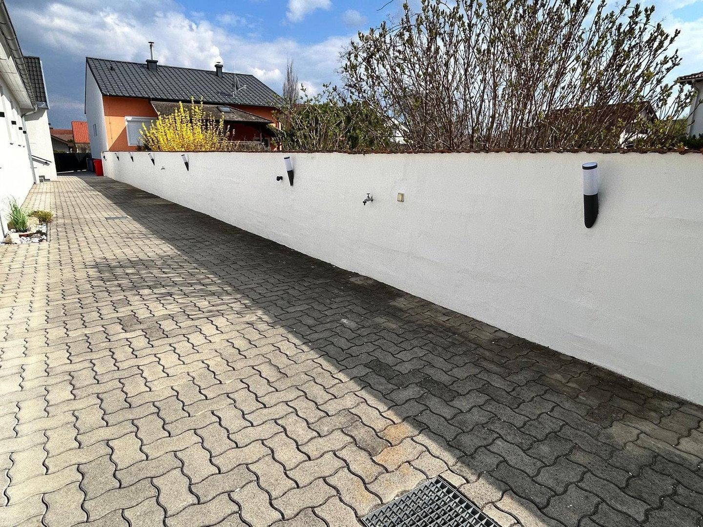 Gepflasterte Zufahrt mit einer langen weißen Mauer und Außenbeleuchtung.