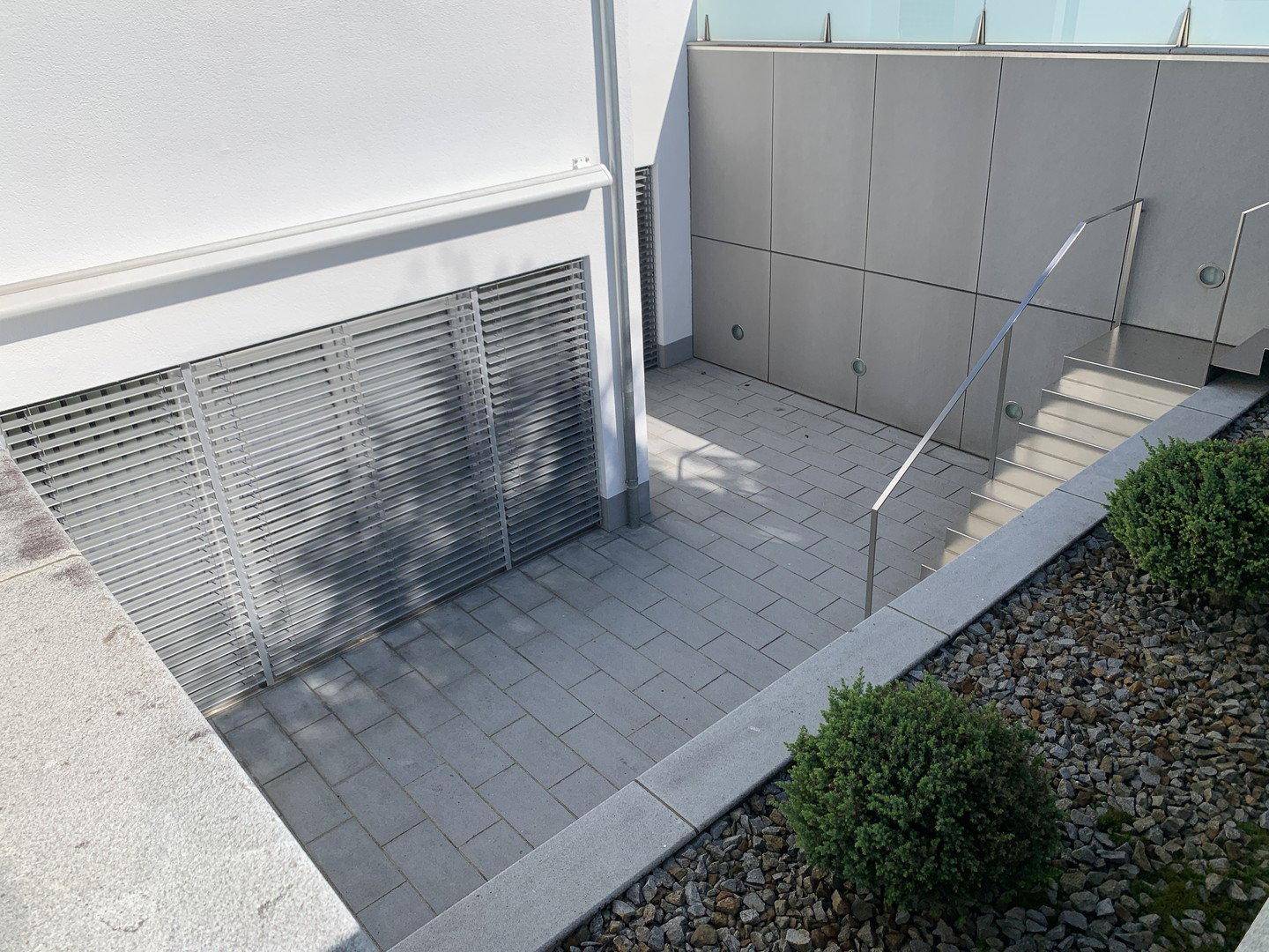 Zugang zum Untergeschoss mit Außentreppe und gepflastertem Bereich, umgeben von kleinen Sträuchern.