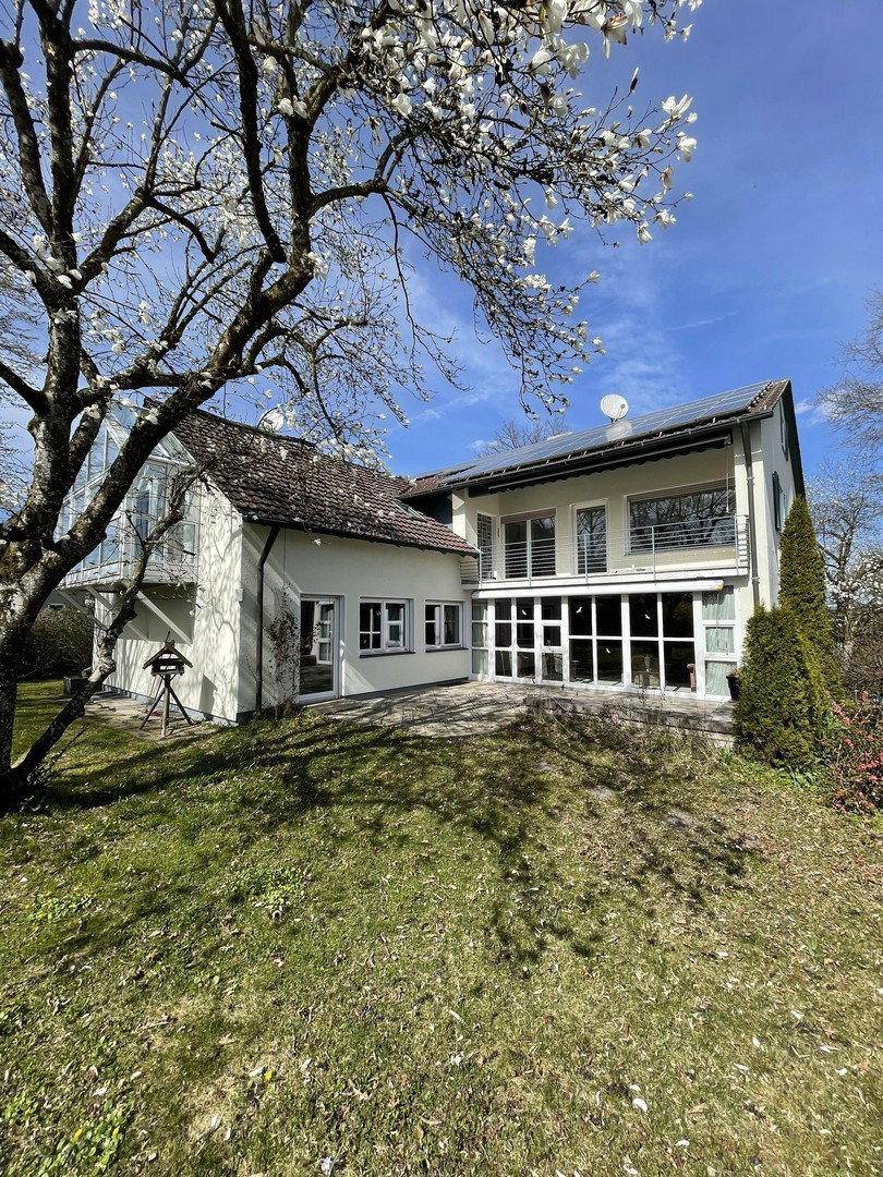 Hinteransicht des Hauses mit Terrasse, Garten und blühenden Bäumen im Frühling.