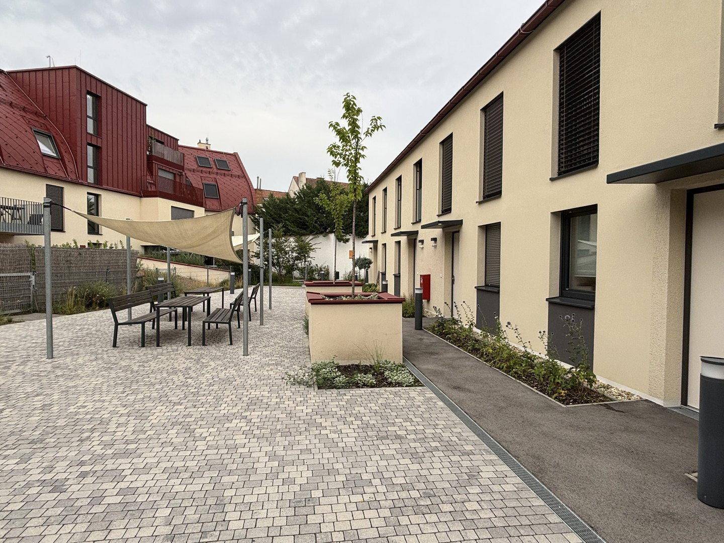 Wohnanlage mit heller Fassade, gepflastertem Innenhof und einem schattigen Sitzbereich im Freien.