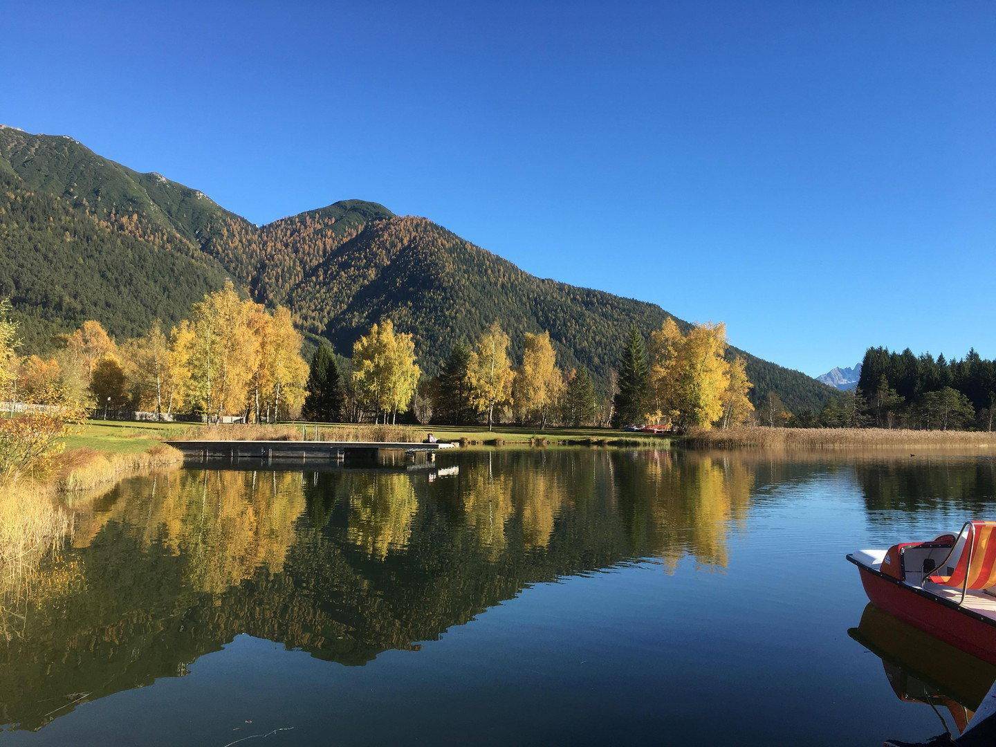 Idyllischer See umgeben von herbstlichen Bäumen und Bergen, mit einem kleinen Boot am Ufer.