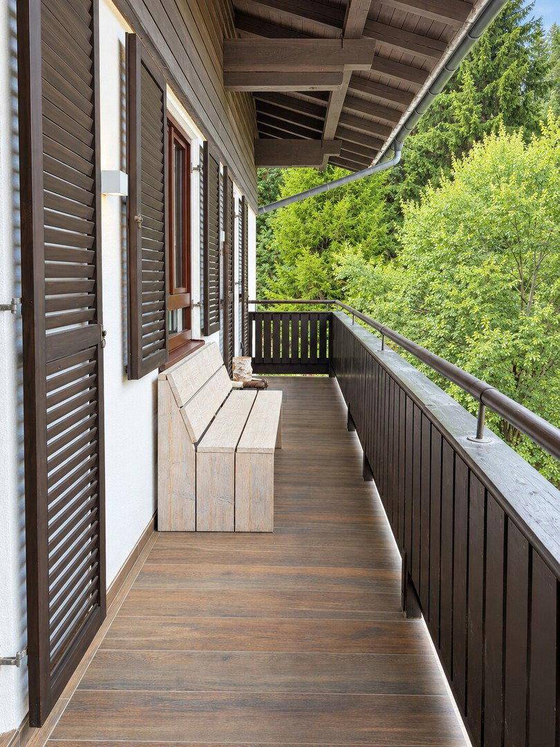 Langer Balkon mit Holzboden und Sitzbank, umgeben von grünen Bäumen, ideal zum Entspannen.