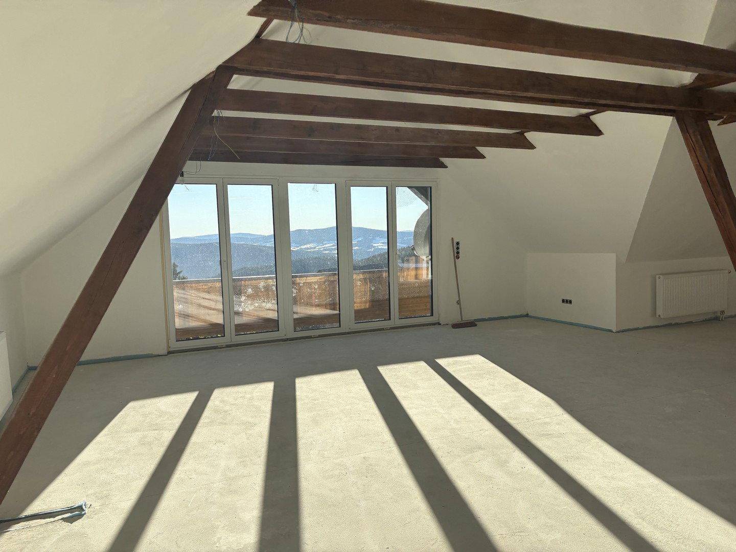 Heller Dachbodenraum mit Holzbalken, großen Fenstern und weitem Bergblick, noch im Ausbau.