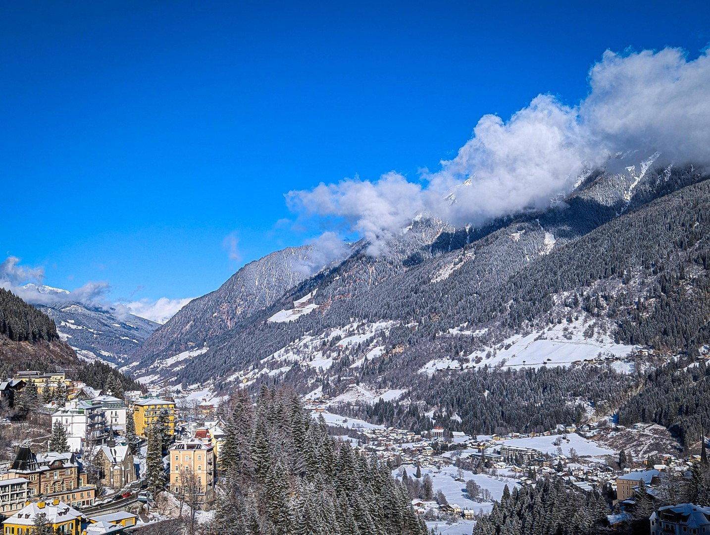 Winterlicher Panoramablick auf ein verschneites Tal mit einem Dorf, umgeben von schneebedeckten Wäldern und Bergen.