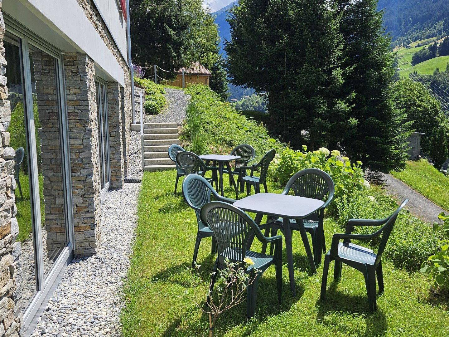 Gemütlicher Sitzbereich im Garten mit Tischen und Stühlen, umgeben von Grün und Bergblick.