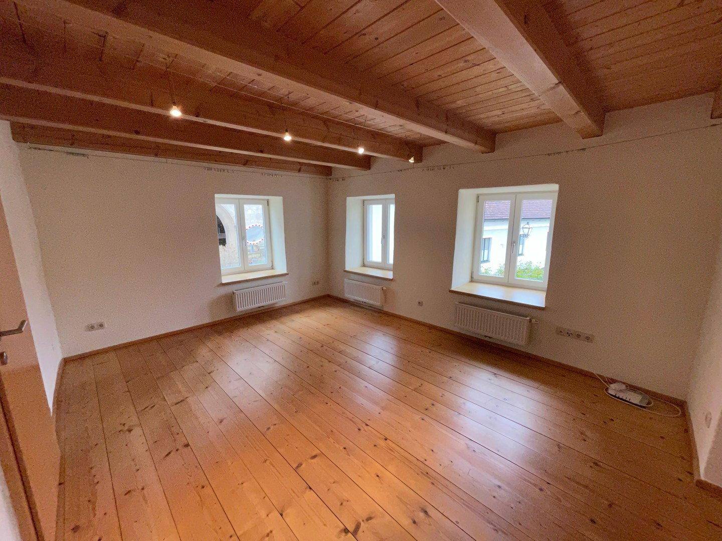 Geräumiger Wohnbereich mit warmem Holzdielenboden und rustikalen Deckenbalken, gut beleuchtet.