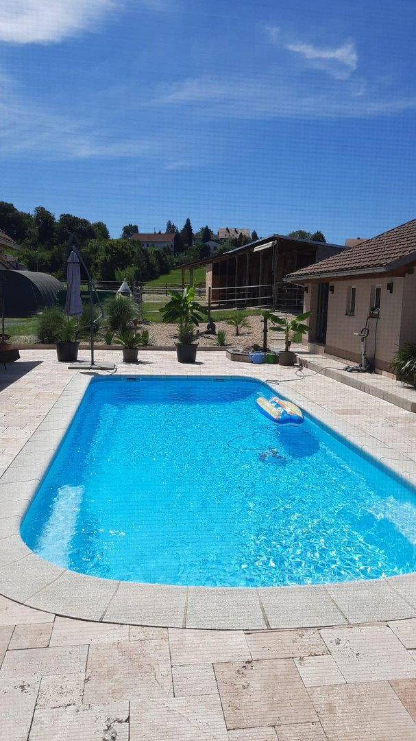 Großer Swimmingpool mit blauer Wasseroberfläche, umgeben von einer hellen Steinterrasse und grüner Landschaft.
