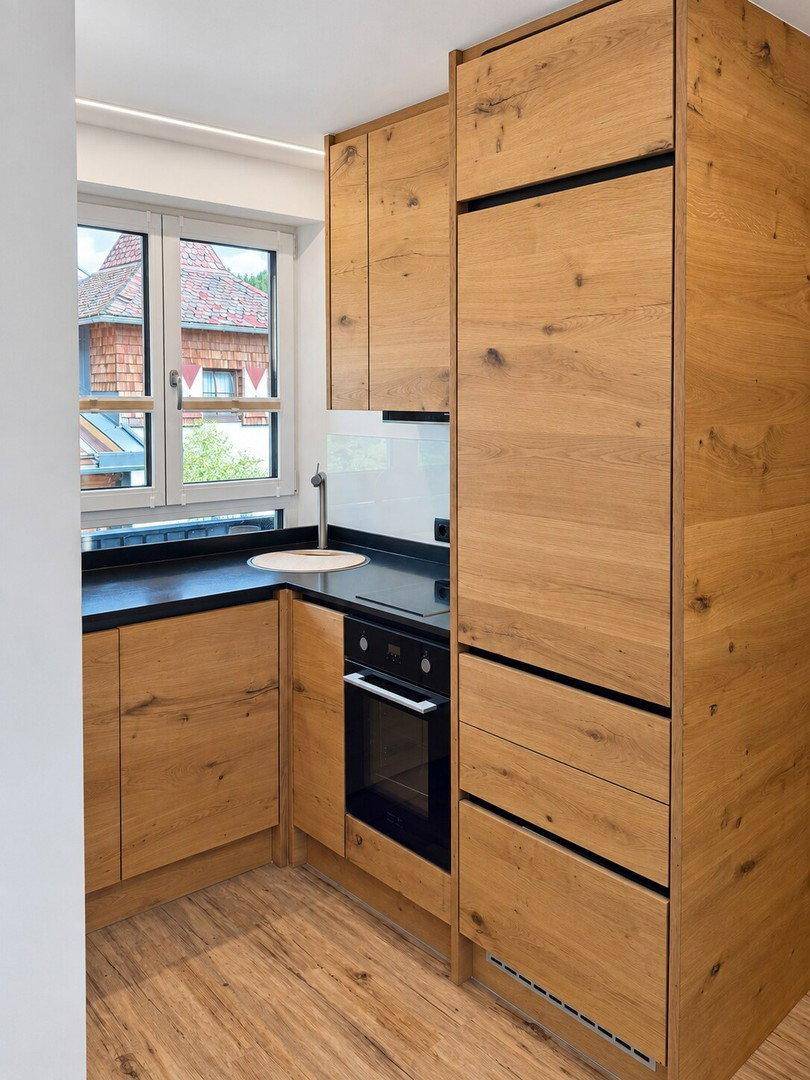 Kompakte Küche mit Holzfronten, schwarzer Arbeitsplatte und integrierten Geräten unter dem Fenster.