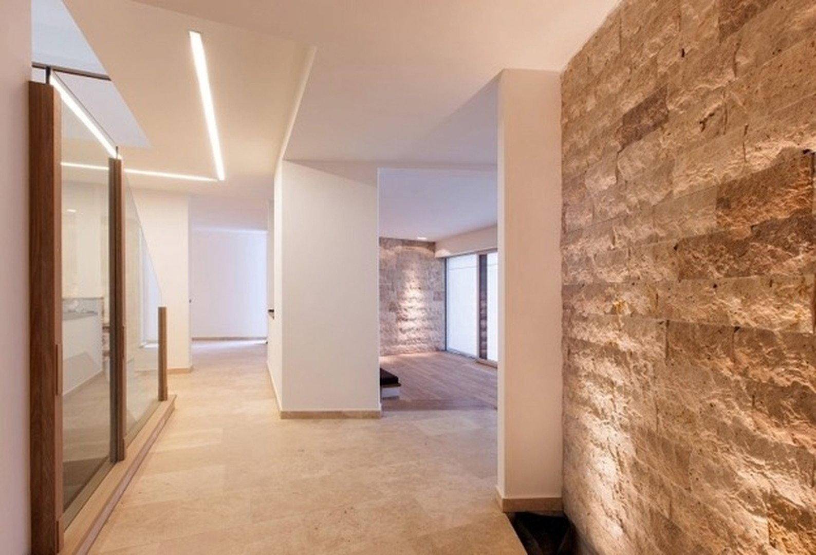 Heller Flur mit Natursteinwand, modernen Lichtleisten und Glasgeländer zur Treppe.