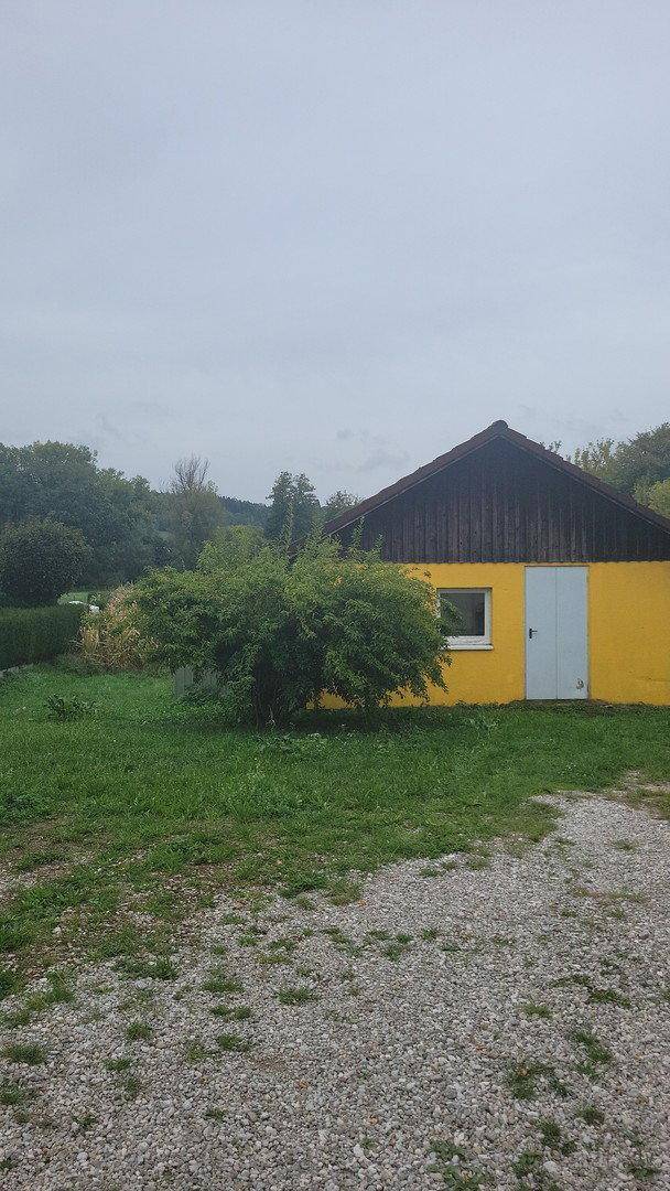 Kleines gelbes Nebengebäude mit dunklem Dach, umgeben von einer Grünfläche und Kies.