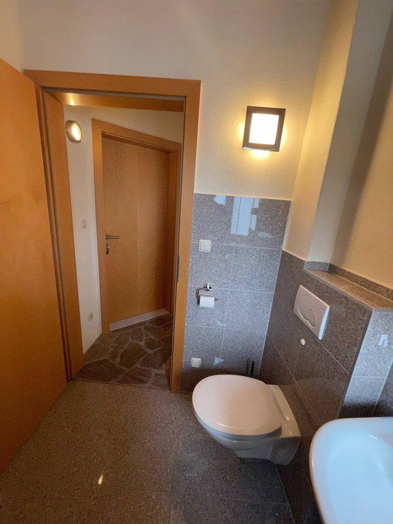 Badezimmer mit grauen Fliesen, wandhängendem WC und Zugang zu einem weiteren Raum.