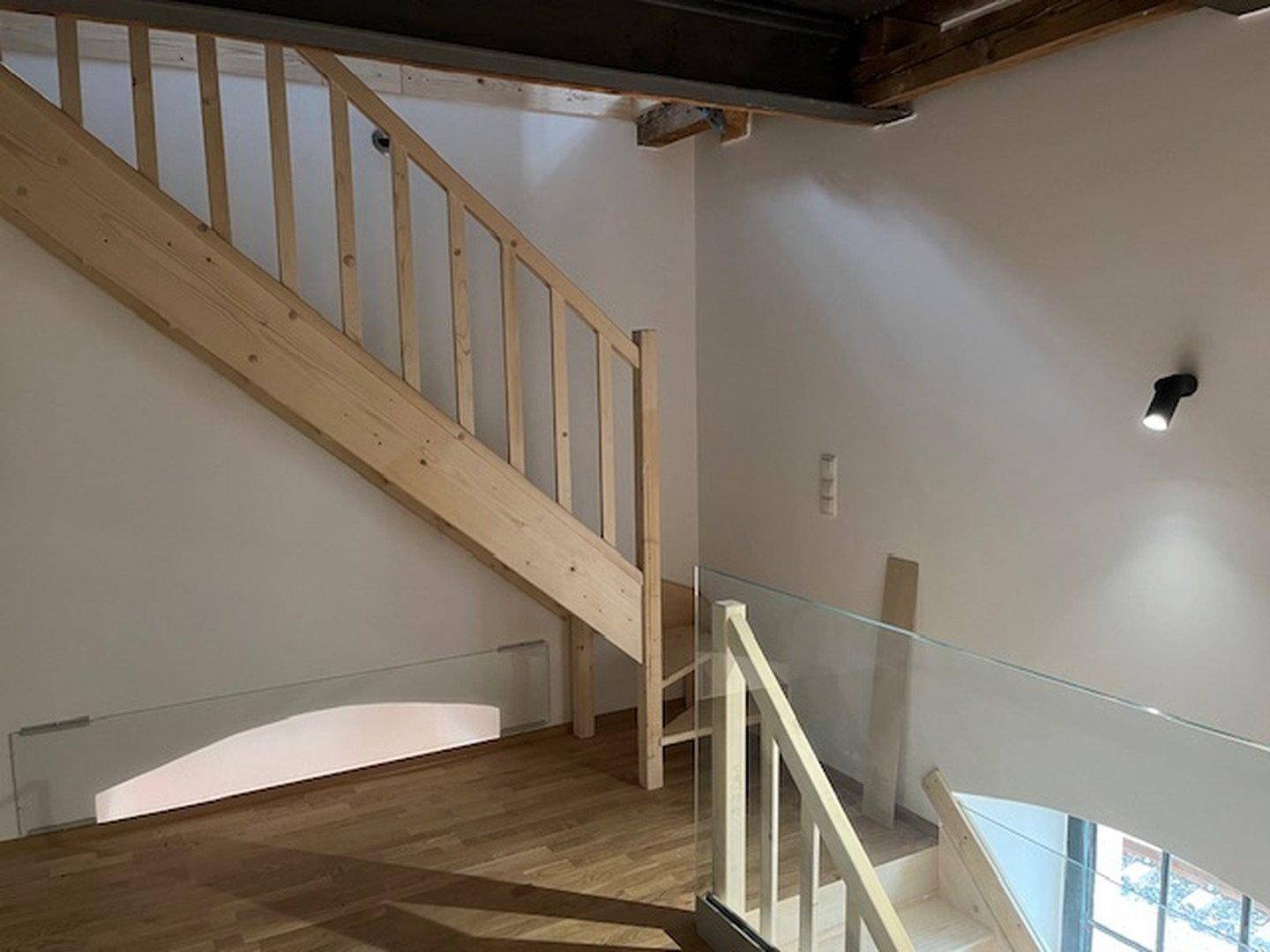 Treppenaufgang mit Holzstufen und Glasgeländer, führt zu einem hellen Raum mit Holzbalken.