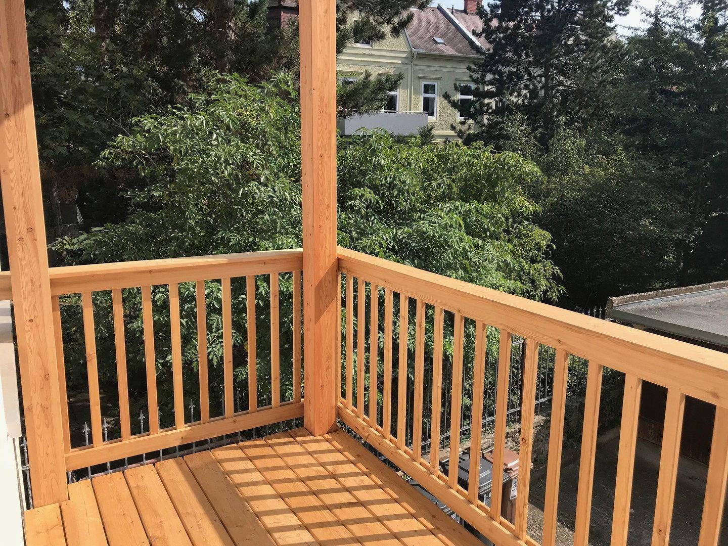 Balkon mit Holzgeländer und Blick auf die grüne Umgebung mit Bäumen und weiteren Gebäuden.