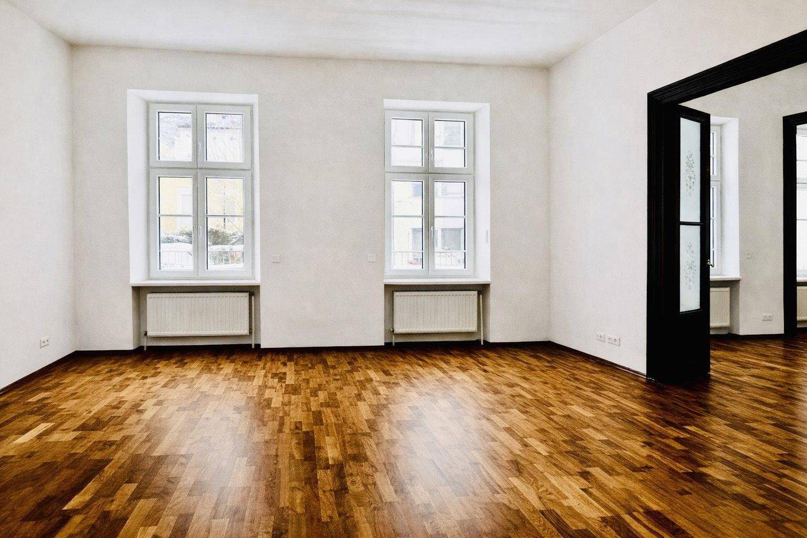 Helles Wohnzimmer mit glänzendem Parkettboden, hohen Decken und zwei großen Fenstern.