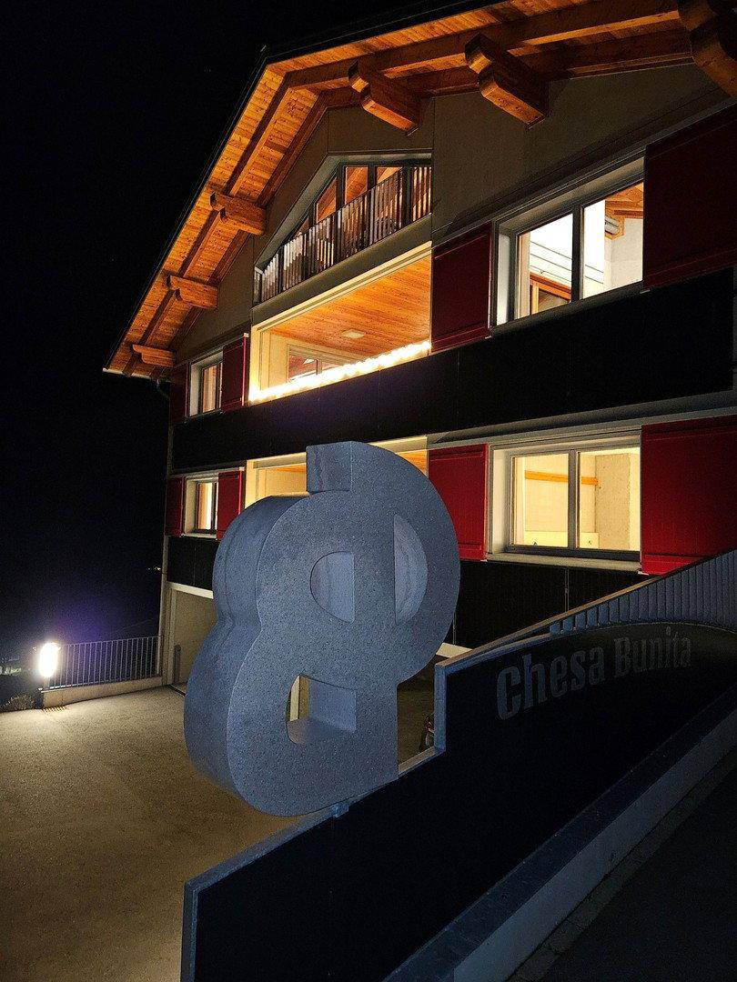 Beleuchtetes Gebäude bei Nacht mit markanter Skulptur und dem Schriftzug "Chesa Bunita" im Vordergrund.
