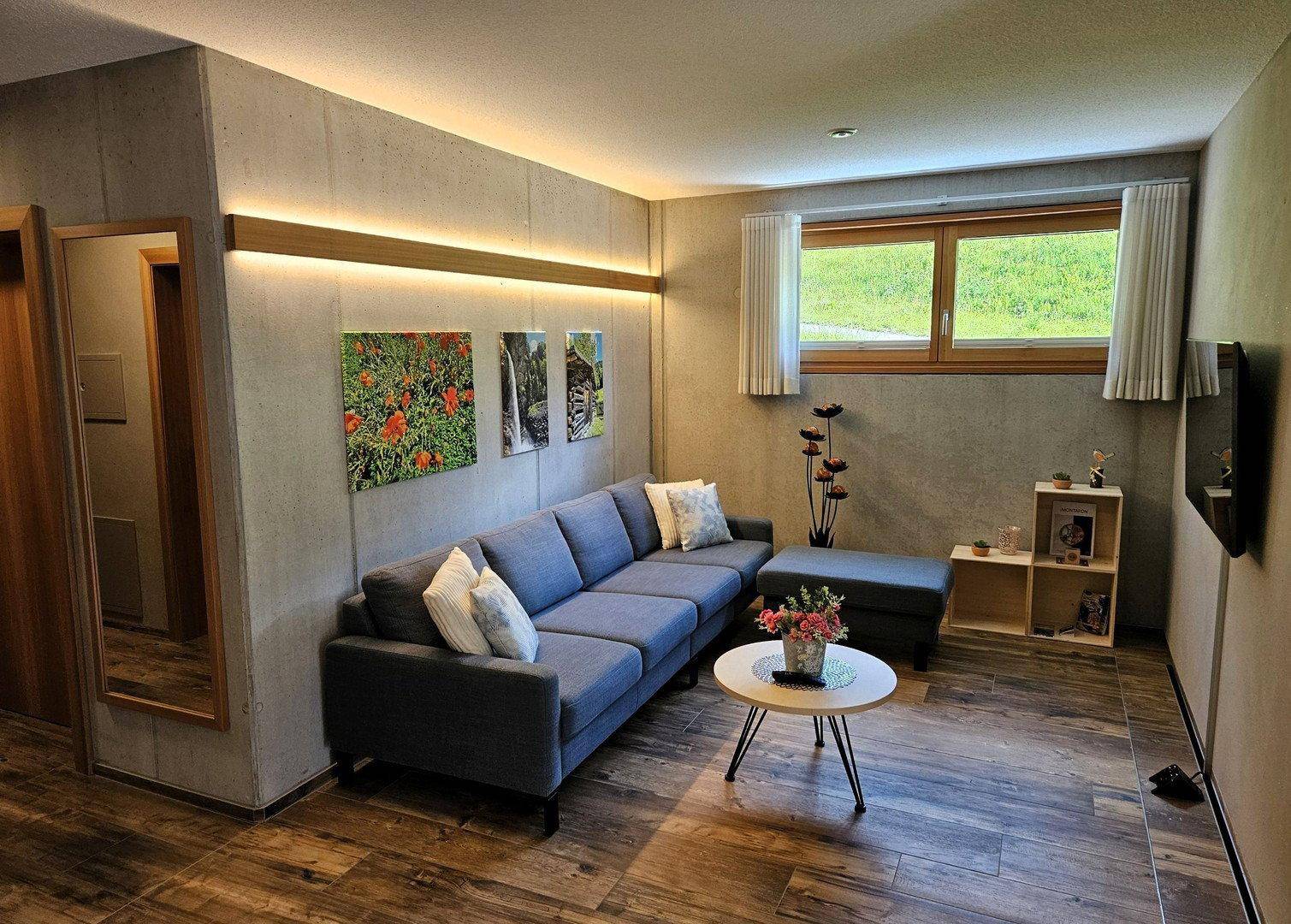 Helles Wohnzimmer mit Holzboden, modernem Sofa und Fenstern mit Blick ins Grüne.