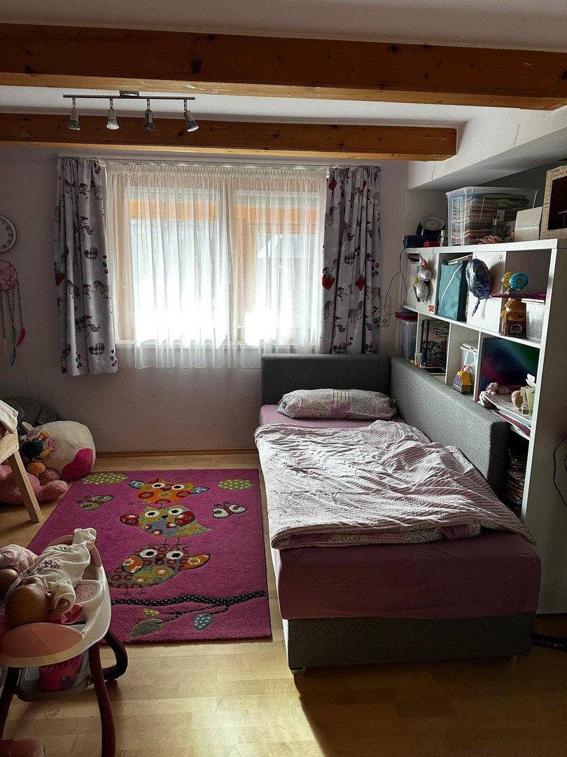 Kinderzimmer mit Einzelbett, Holzbalken und Fenster mit Vorhängen, hell und freundlich.