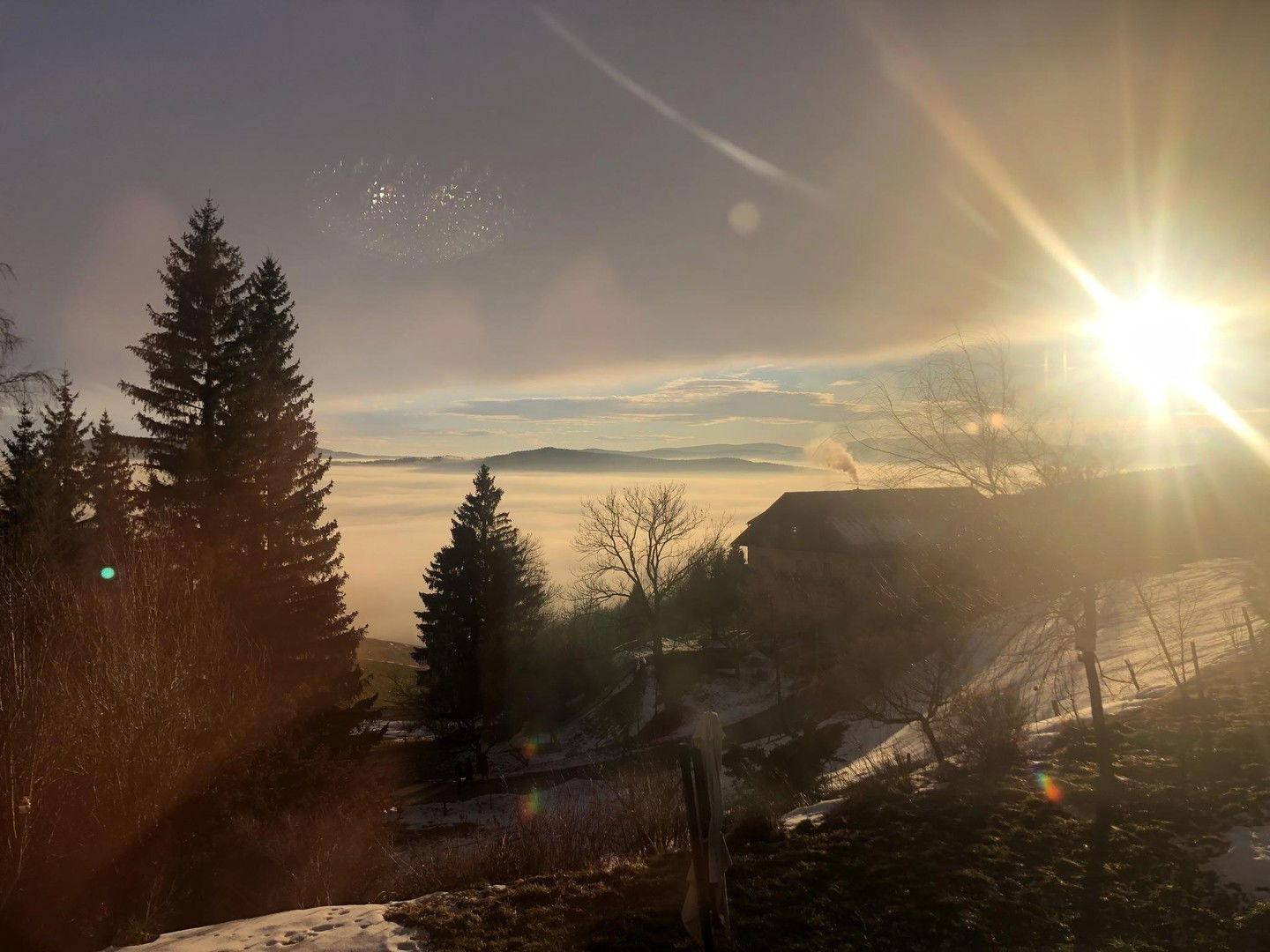 Sonnenaufgang über einer nebligen Berglandschaft mit einem Haus im Hintergrund.