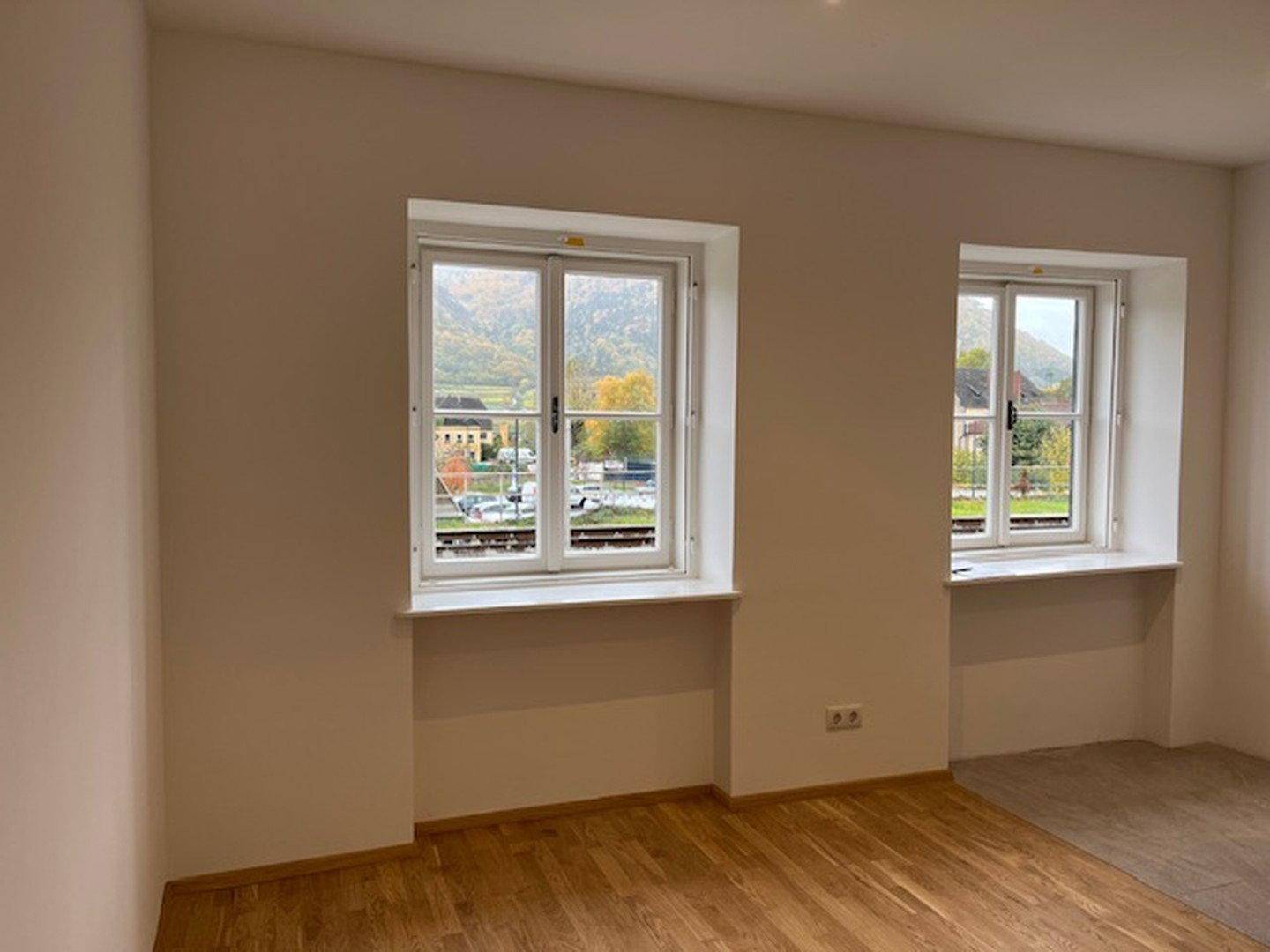 Heller, leerer Raum mit zwei Fenstern, Holzboden und Blick auf die umliegende Landschaft.