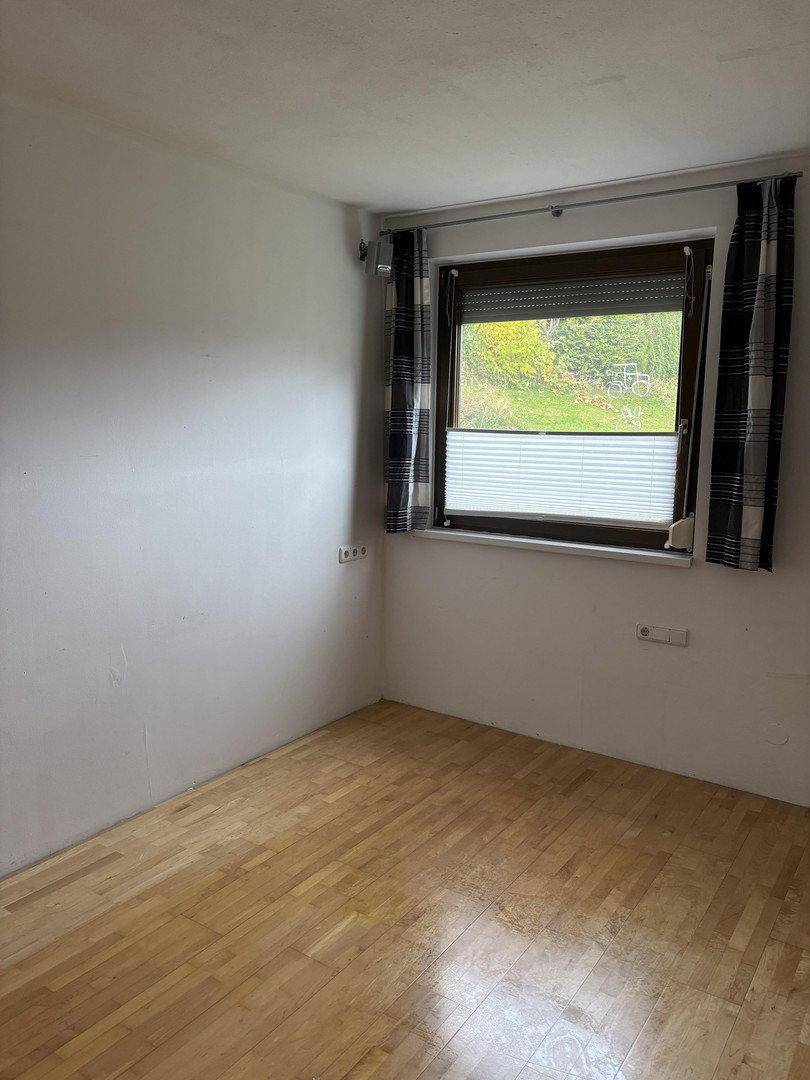 Leerer Raum mit hellem Parkettboden, weißen Wänden und einem Fenster mit Jalousien.