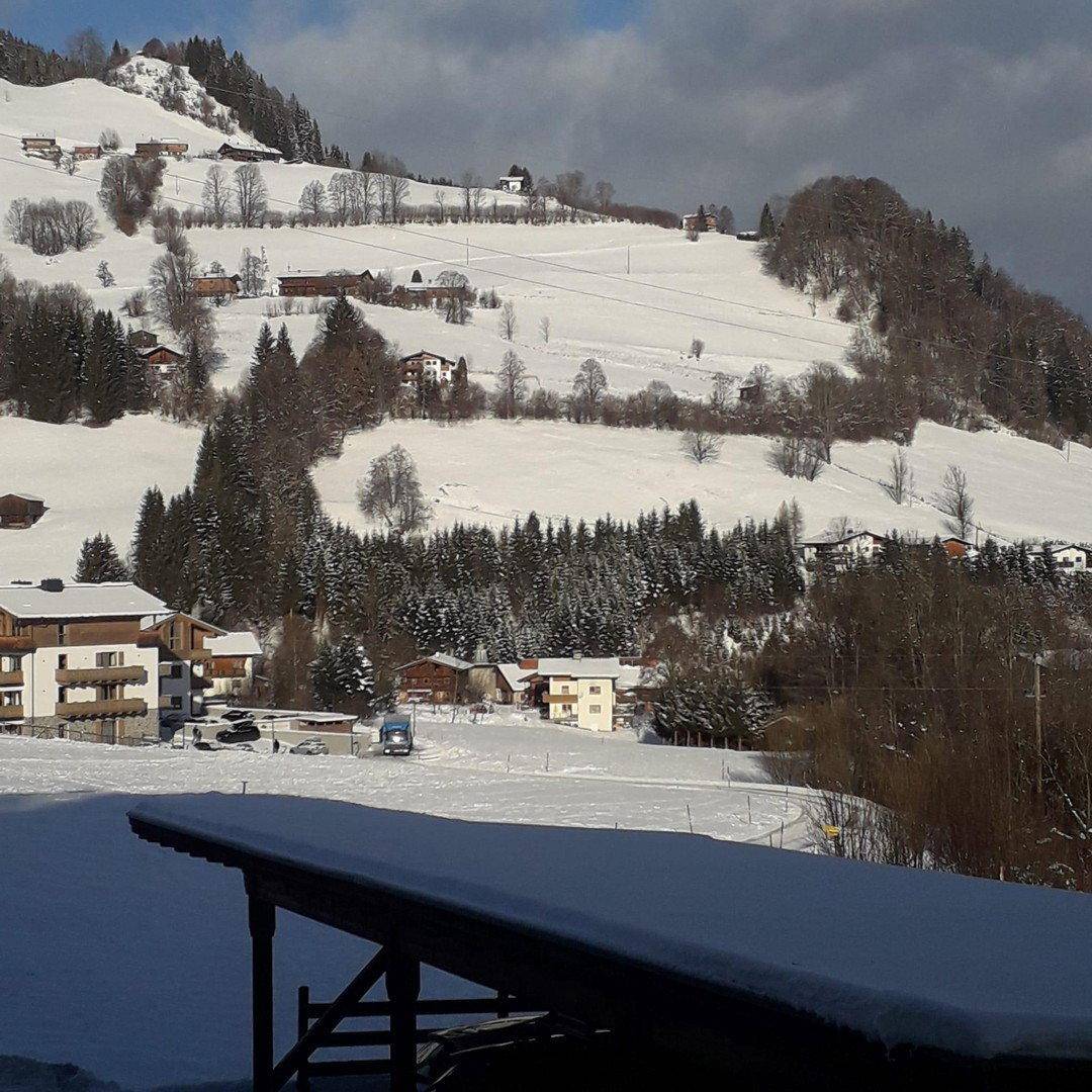 Weitläufige, schneebedeckte Berglandschaft mit verstreuten Häusern und bewaldeten Hängen unter blauem Himmel.