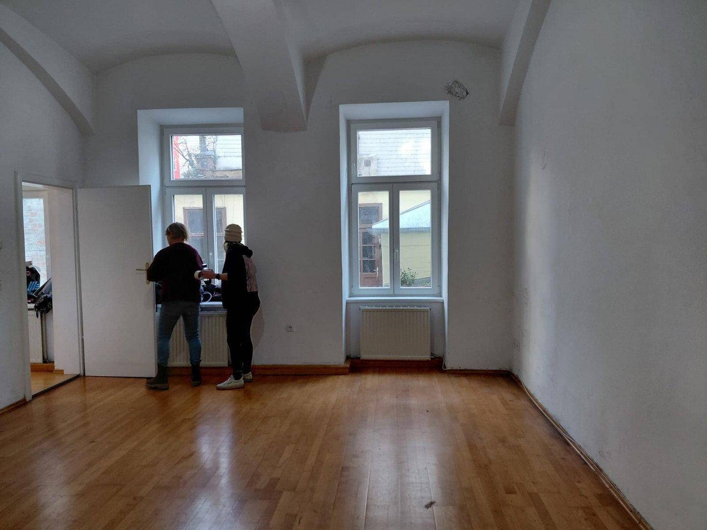 Heller Wohnraum mit Holzboden, zwei Fenstern und Personen, die den Raum besichtigen.