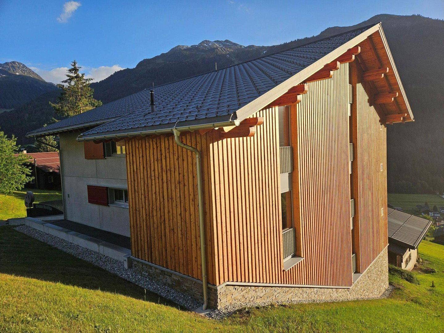 Seitenansicht des Gebäudes mit Holzverkleidung und Satteldach, eingebettet in eine grüne Hügellandschaft.