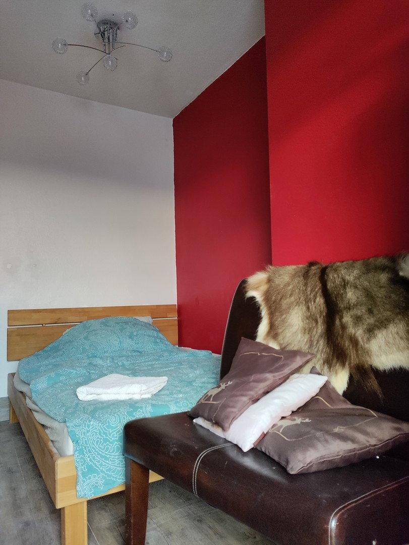 Schlafzimmer mit Holzbett, roter Akzentwand und einer gemütlichen Sitzbank mit Fellauflagen.
