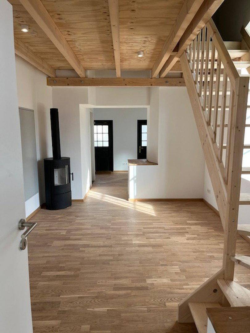 Offener Wohnbereich mit Holzboden, sichtbaren Deckenbalken und modernem Kaminofen.