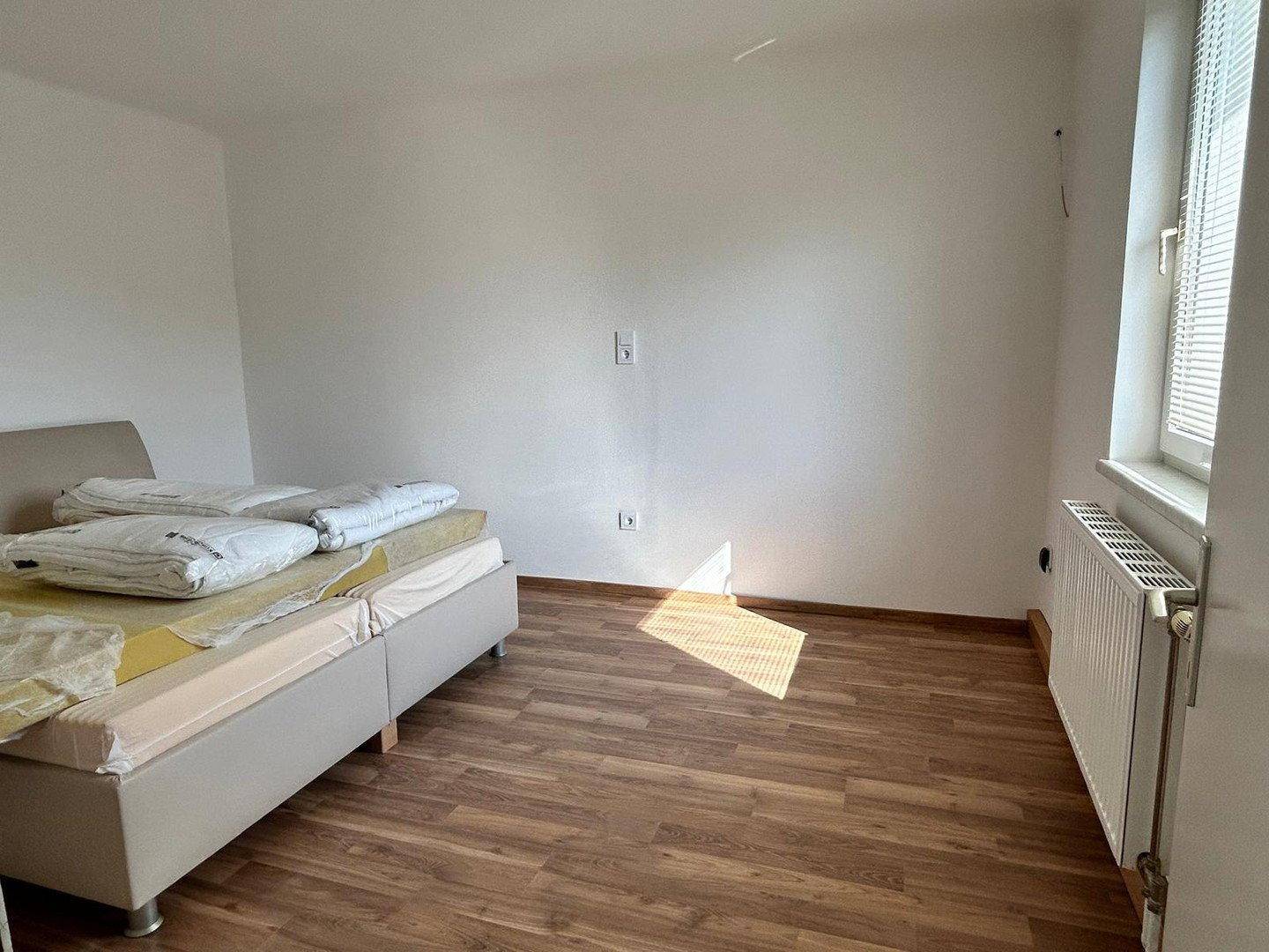 Helles Schlafzimmer mit Laminatboden, Heizkörper und Fenster mit Blick nach draußen.