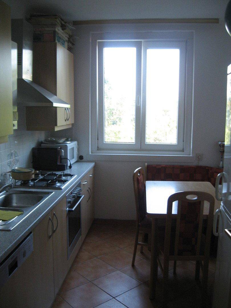 Funktionale Küche mit hellen Holzschränken, Fliesenboden und Essbereich am Fenster.