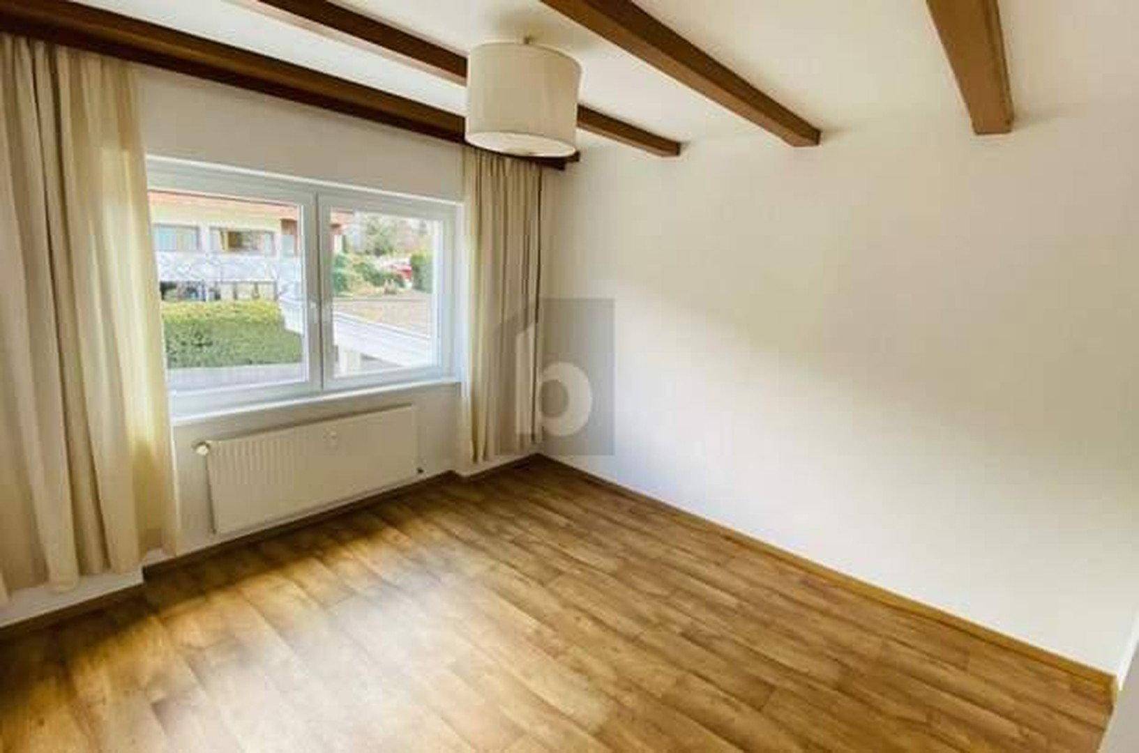 Helles Zimmer mit Holzboden, sichtbaren Deckenbalken und großem Fenster mit Blick ins Grüne.