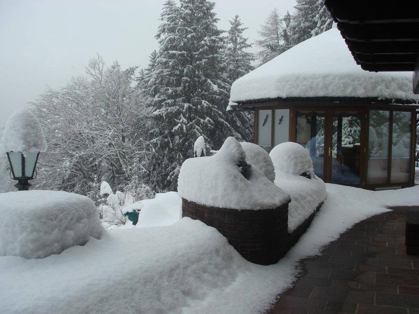 Verschneite Terrasse mit einer Laterne und einem Wintergarten im Hintergrund, umgeben von tiefem Schnee.