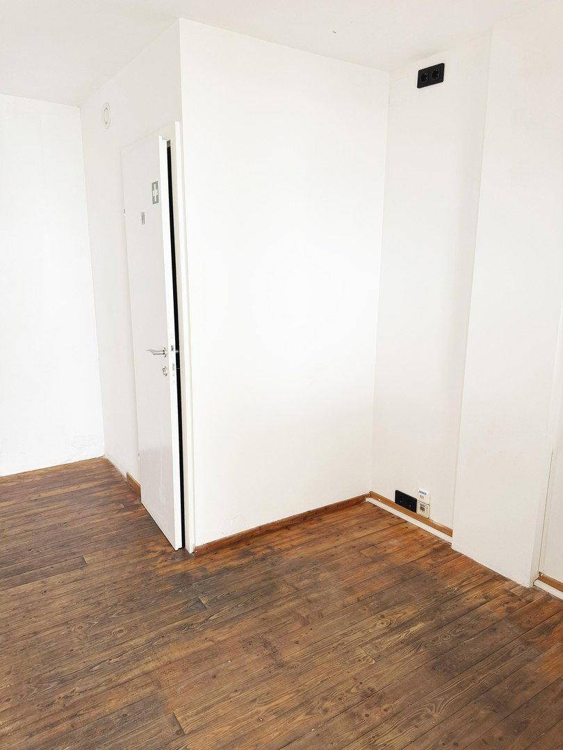 Einfacher Raum mit dunklem Holzfußboden und einer weißen Tür, die zu einem weiteren Bereich führt.
