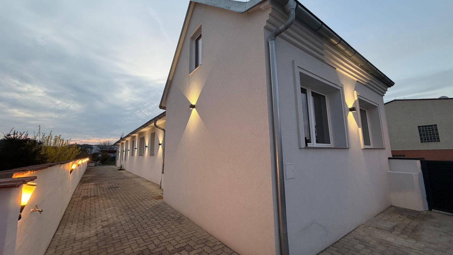 Seitenansicht des Gebäudes bei Dämmerung mit beleuchteter Fassade und gepflastertem Zugang.