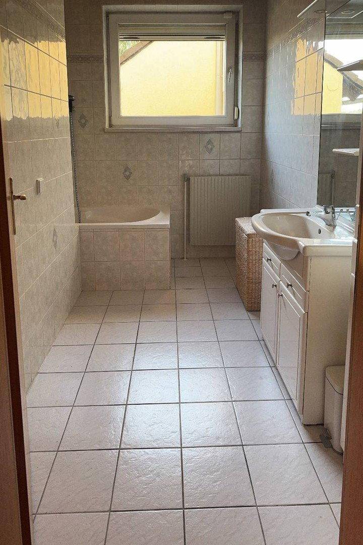 Helles Badezimmer mit beigefarbenen Fliesen, Badewanne und Fenster für Tageslicht.
