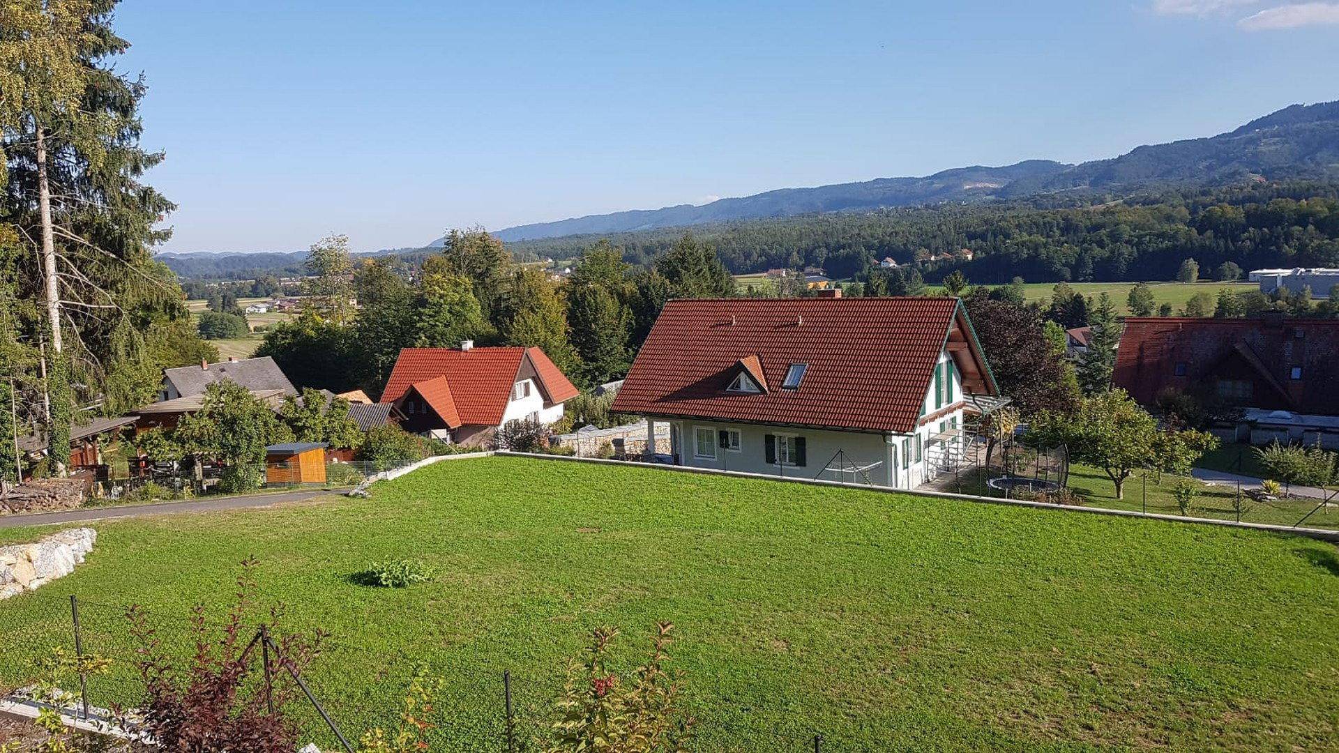 Weitläufiger Blick über grüne Wiesen und Wohnhäuser mit bewaldeten Bergen am Horizont unter blauem Himmel.