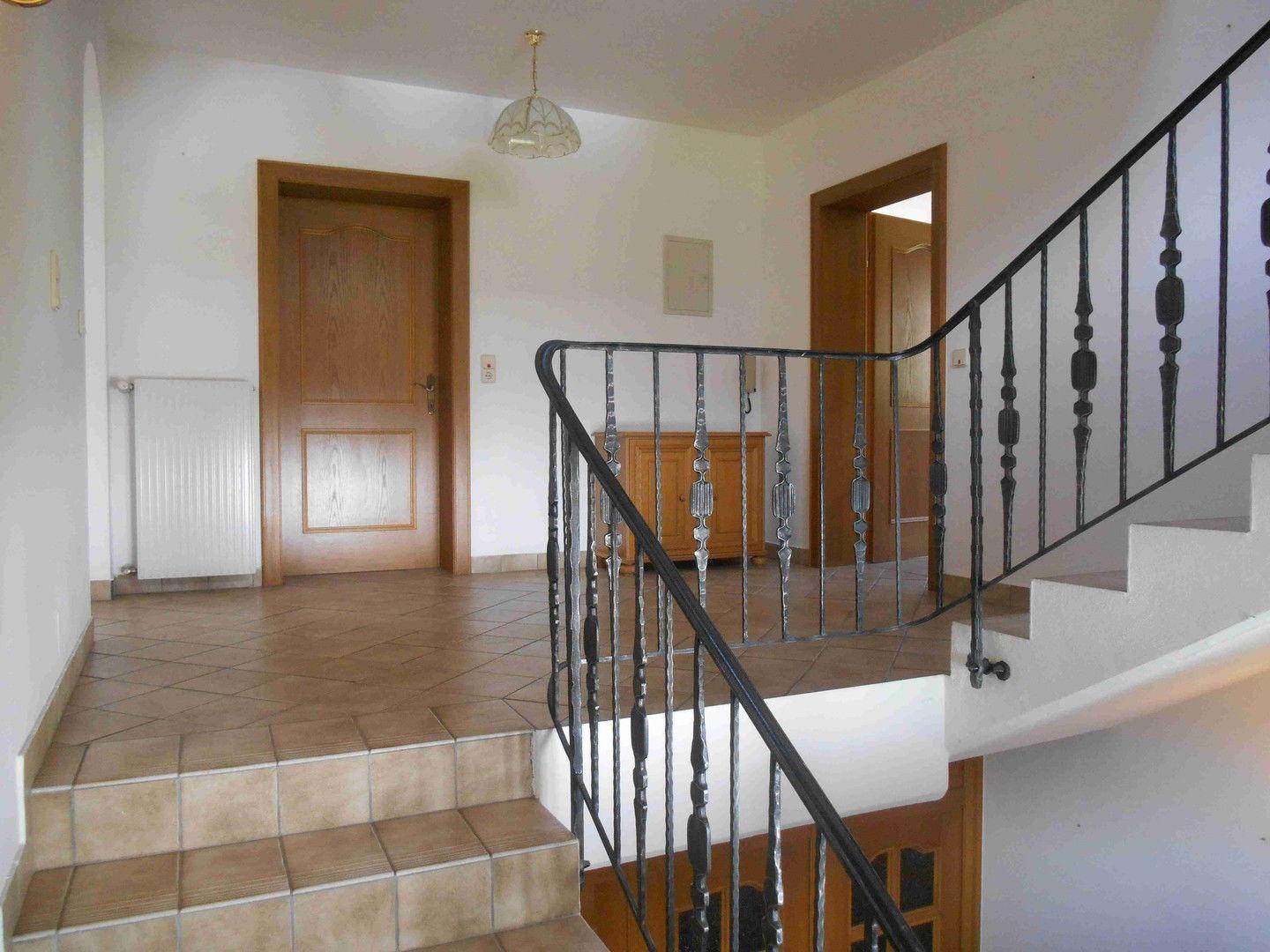 Heller Flur mit gefliestem Boden, schmiedeeisernem Geländer und Holztreppe zum Obergeschoss.