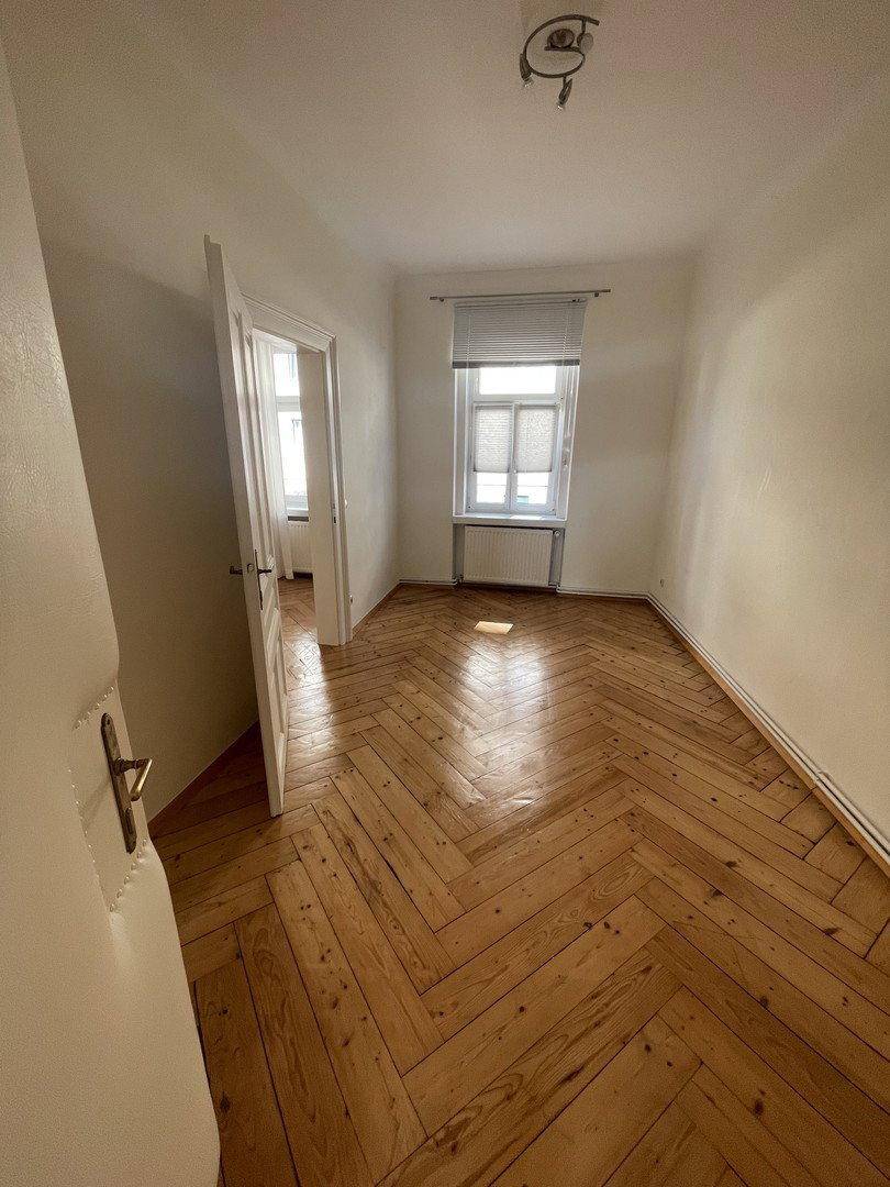 Helles Zimmer mit Fischgrätparkett, weißen Wänden und einem Fenster mit Jalousien.