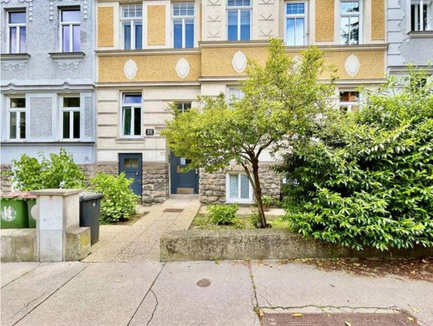 Historisches Gebäude mit gelber Fassade, blauen Türen und grüner Bepflanzung im Vorgarten.