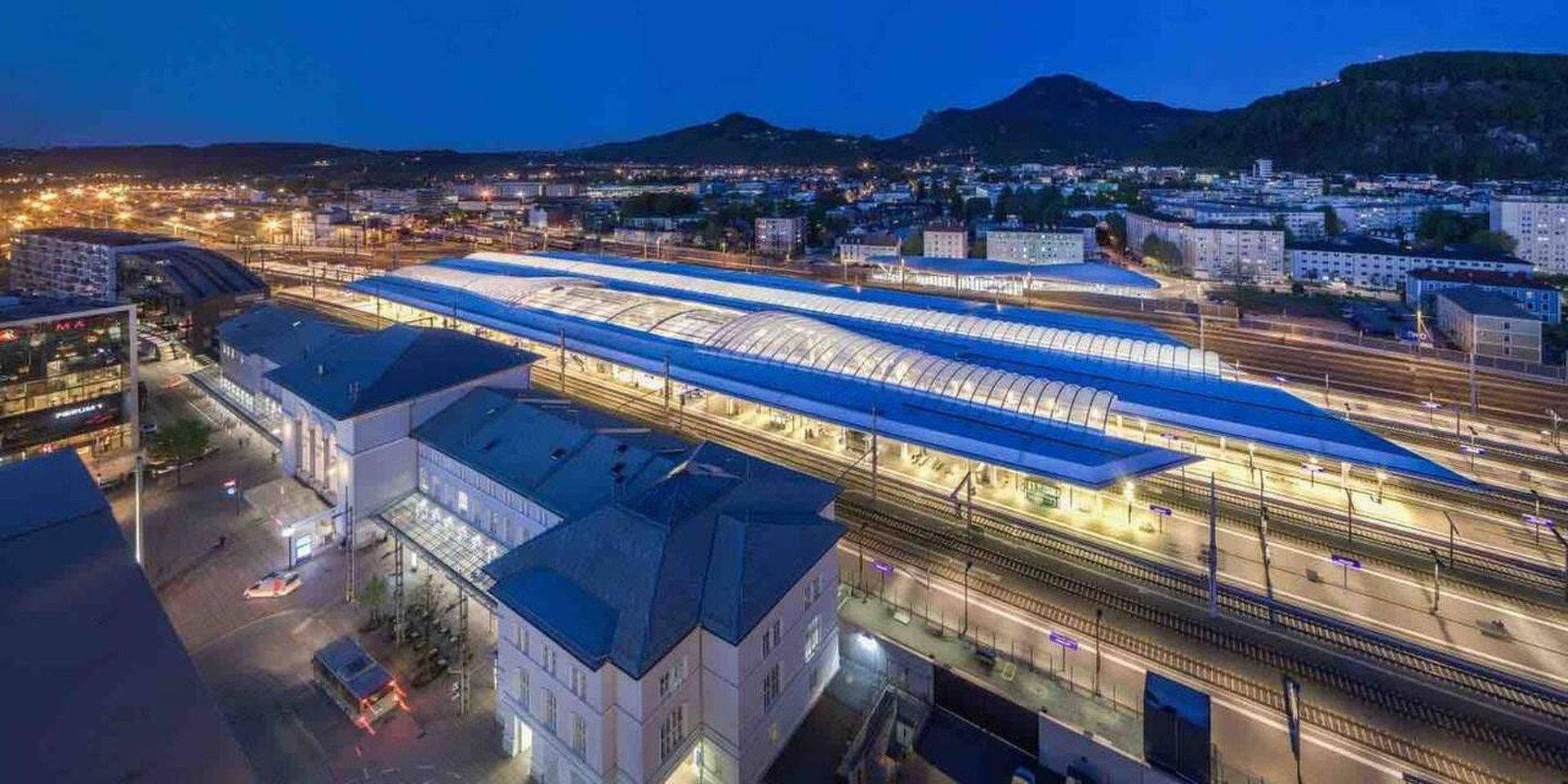 Beeindruckende Nachtansicht eines modernen Bahnhofs mit Stadtlichtern und Bergen im Hintergrund.