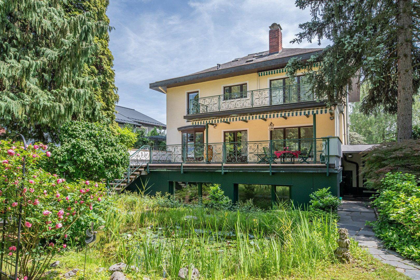 Charmantes Haus mit Balkonen und Terrassen, umgeben von einem grünen Garten mit Teich.