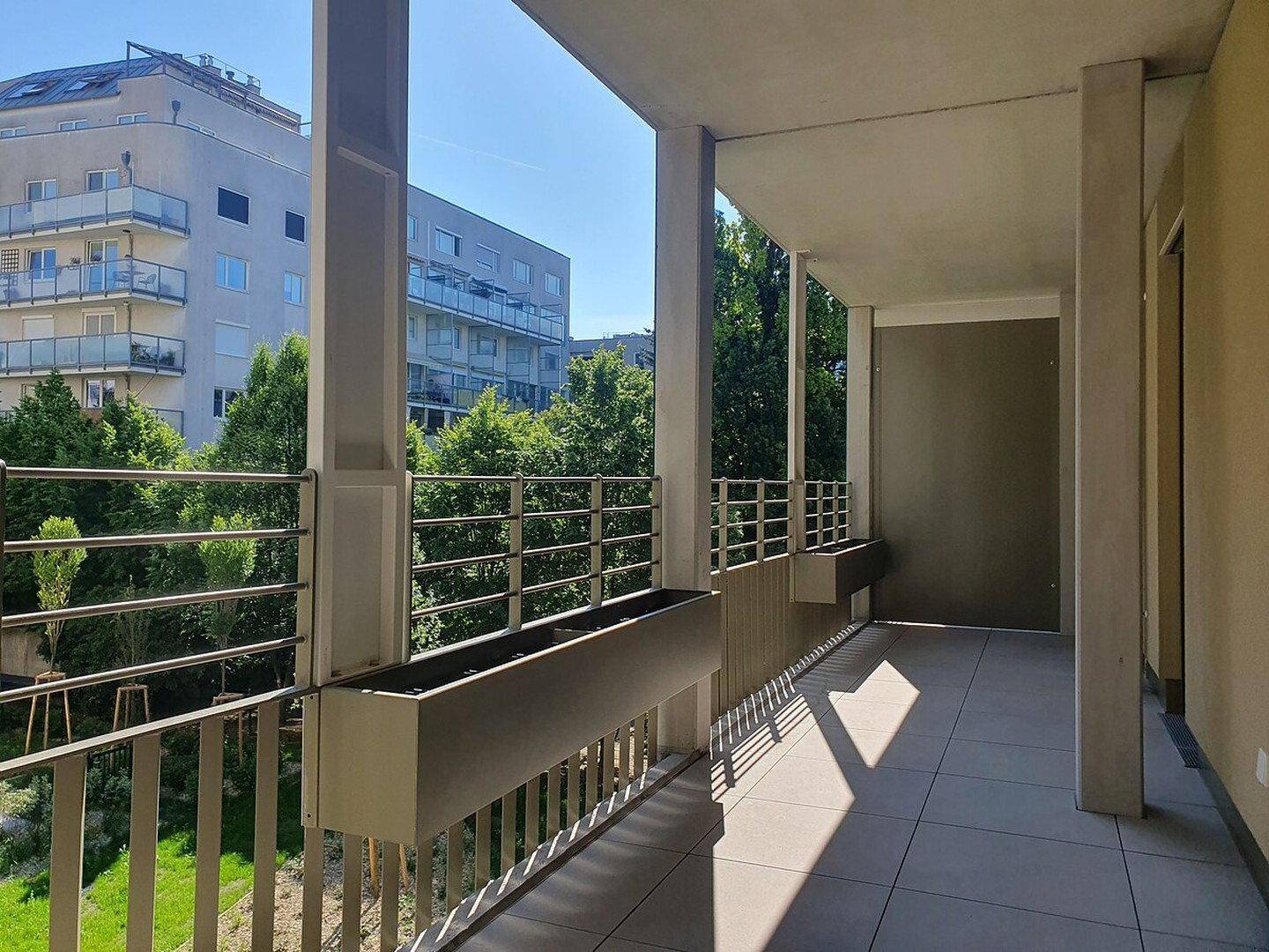 Geräumiger Balkon mit Fliesenboden und Blick auf umliegende Gebäude und grüne Bäume.
