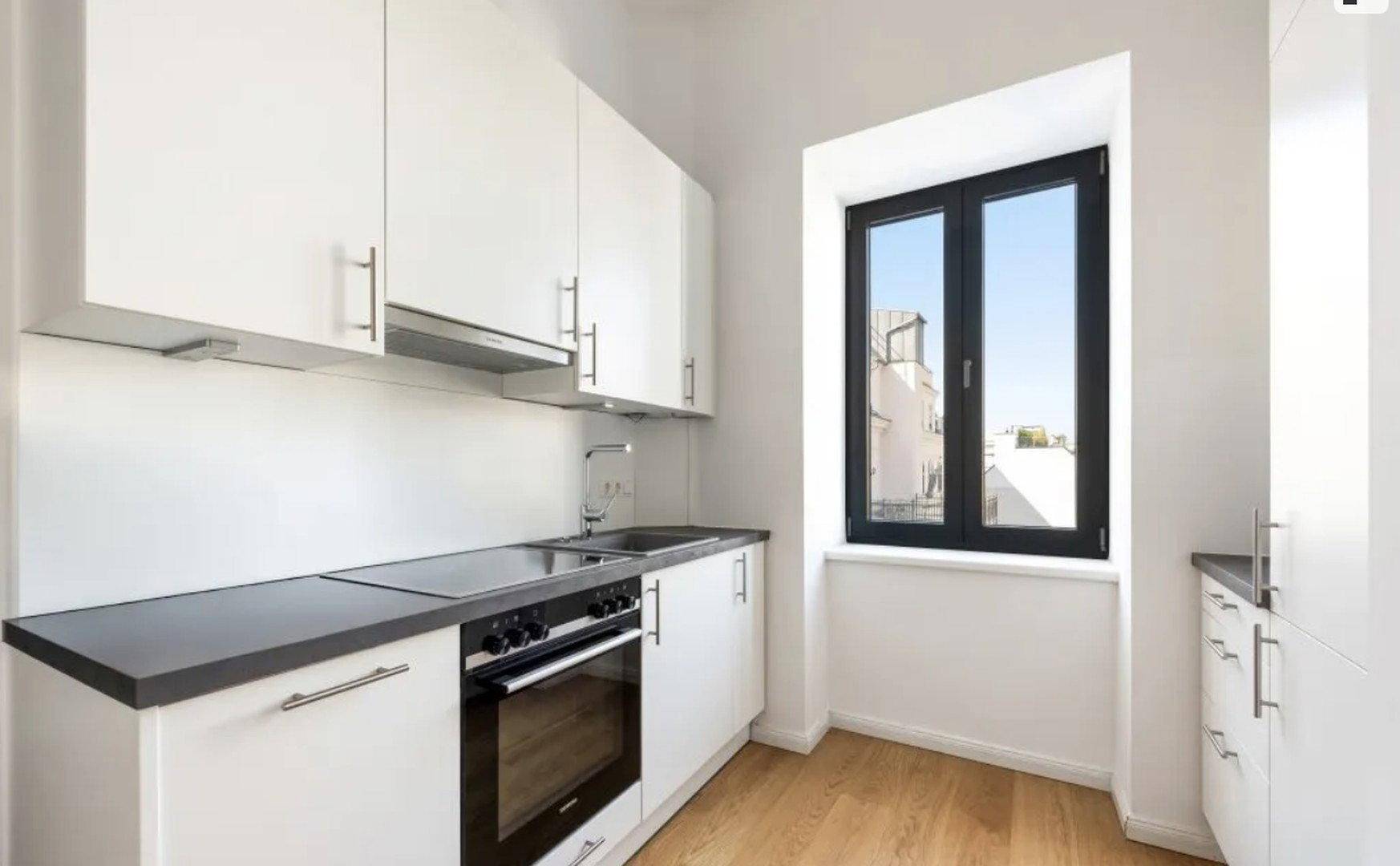 Moderne, kompakte Küche mit weißen Schränken, dunkler Arbeitsplatte und einem Fenster mit Stadtblick.