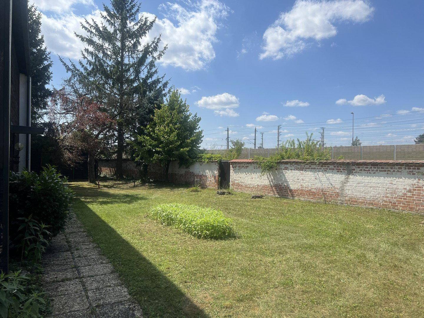 Gartenbereich mit Rasenfläche, alter Ziegelmauer und hohen Bäumen im Hintergrund.