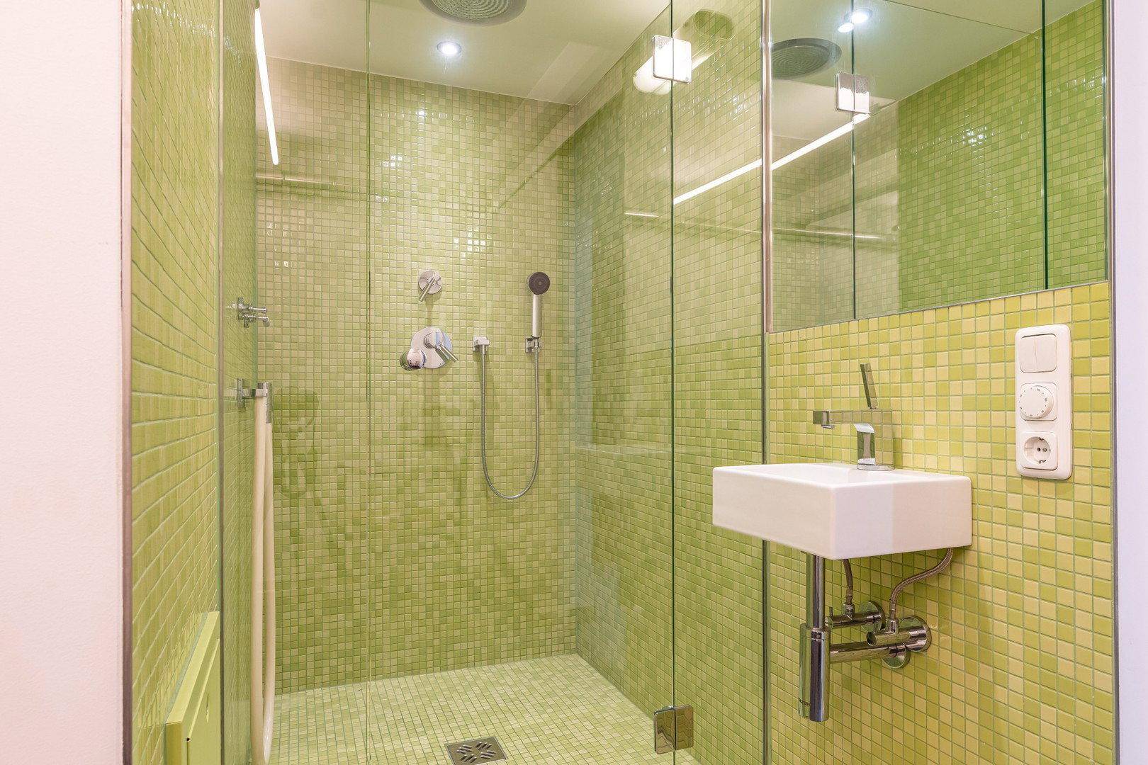 Modernes Badezimmer mit grünen Mosaikfliesen, begehbarer Dusche und kleinem Waschbecken.