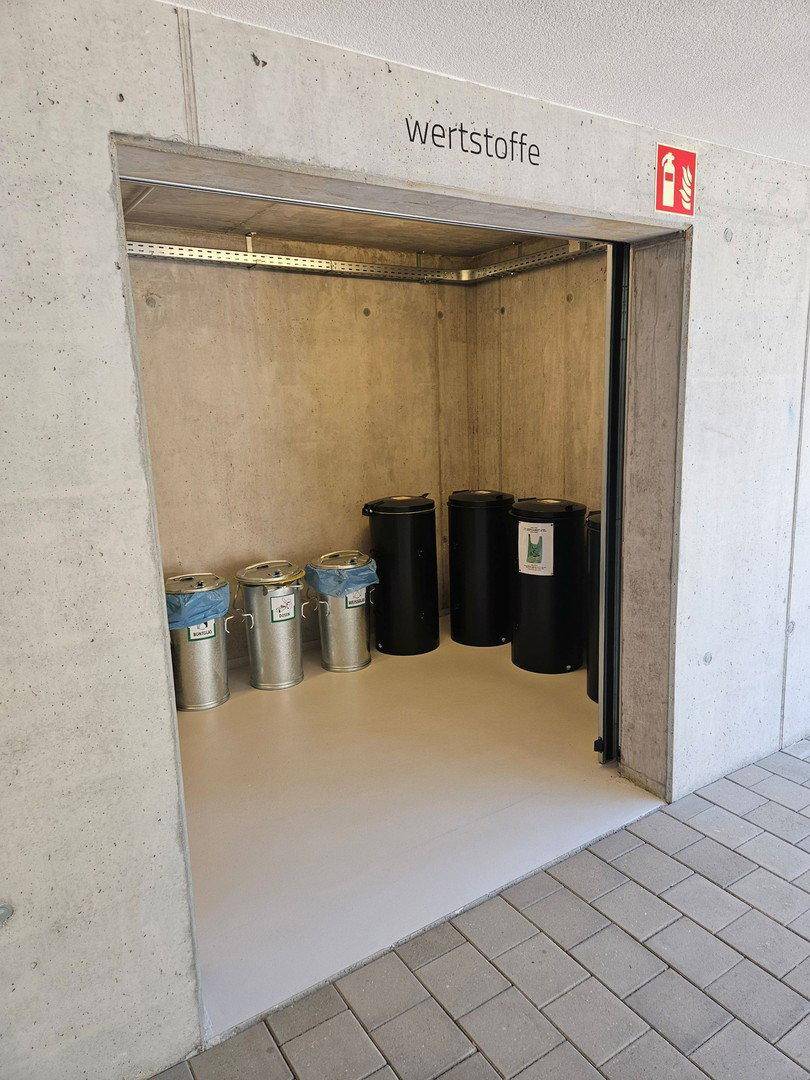 Bereich zur Wertstoffentsorgung mit verschiedenen Mülltonnen und Betonwänden.