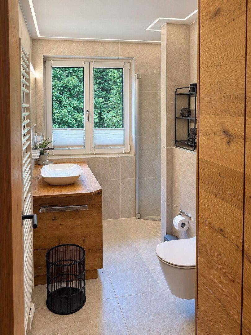 Modernes Badezimmer mit Holzelementen, großformatigen Fliesen und Fenster mit Blick ins Grüne.