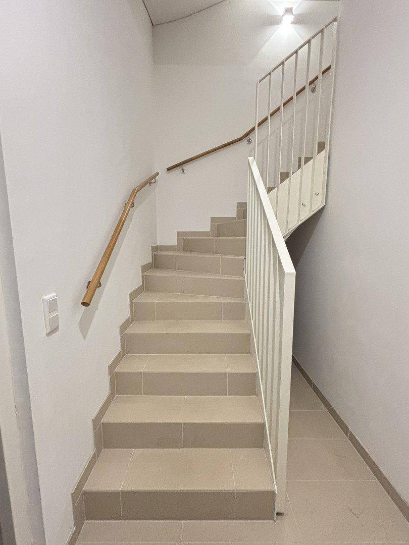 Moderne Treppe mit hellgrauen Fliesenstufen und weißem Geländer, ergänzt durch einen Holzhandlauf.